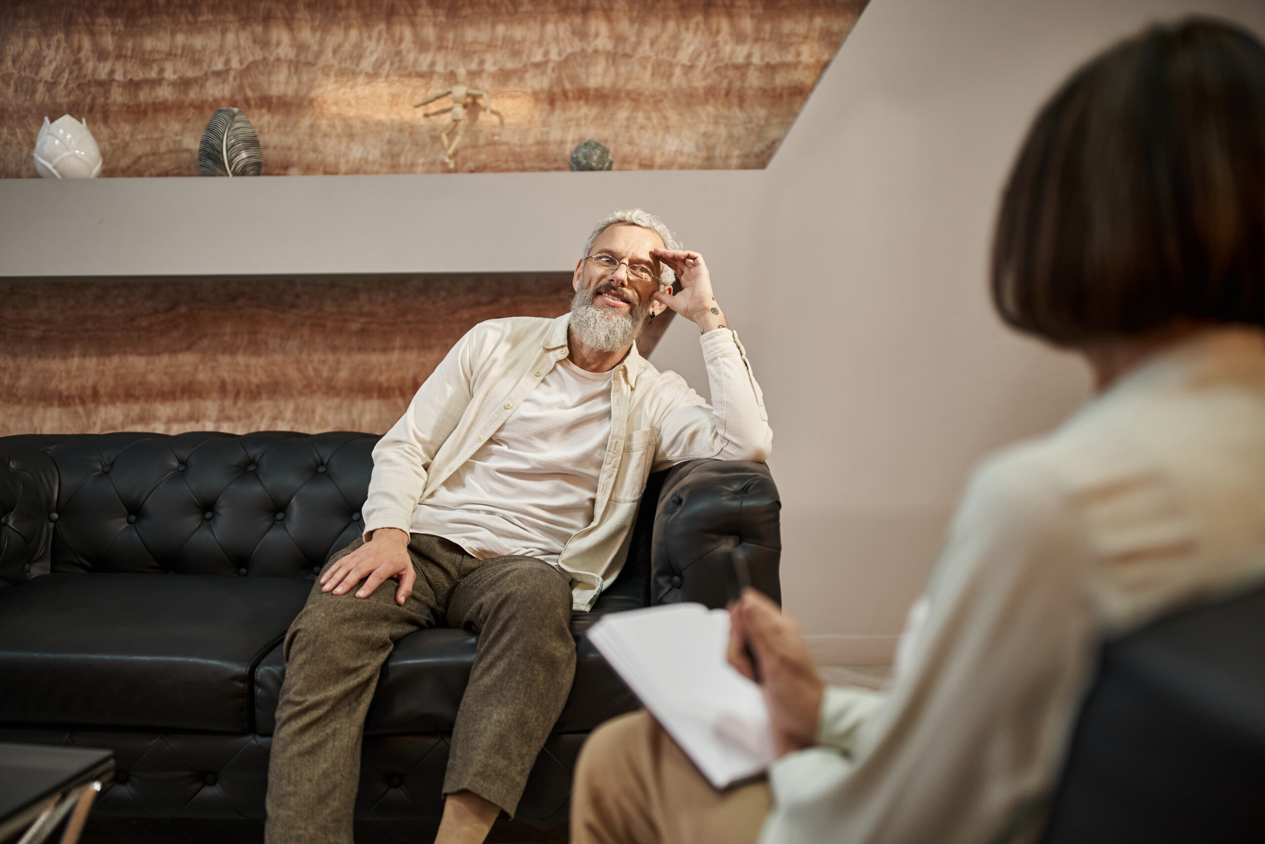 cheerful bearded middled aged man with tattoo sitting on leather couch and looking at psychologist