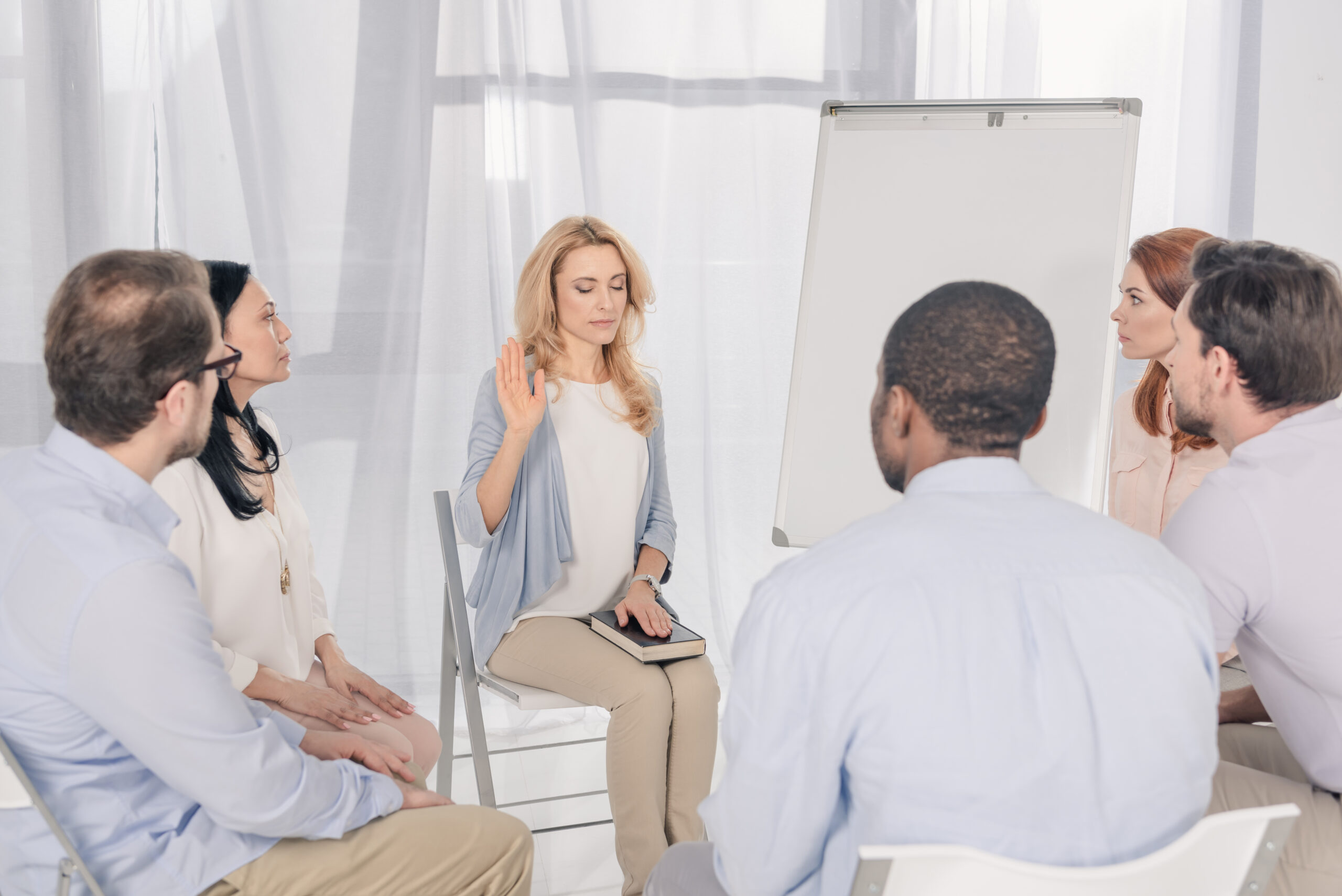 middle aged woman holding hand on Holy Bible while sitting with multiethnic people on chairs at